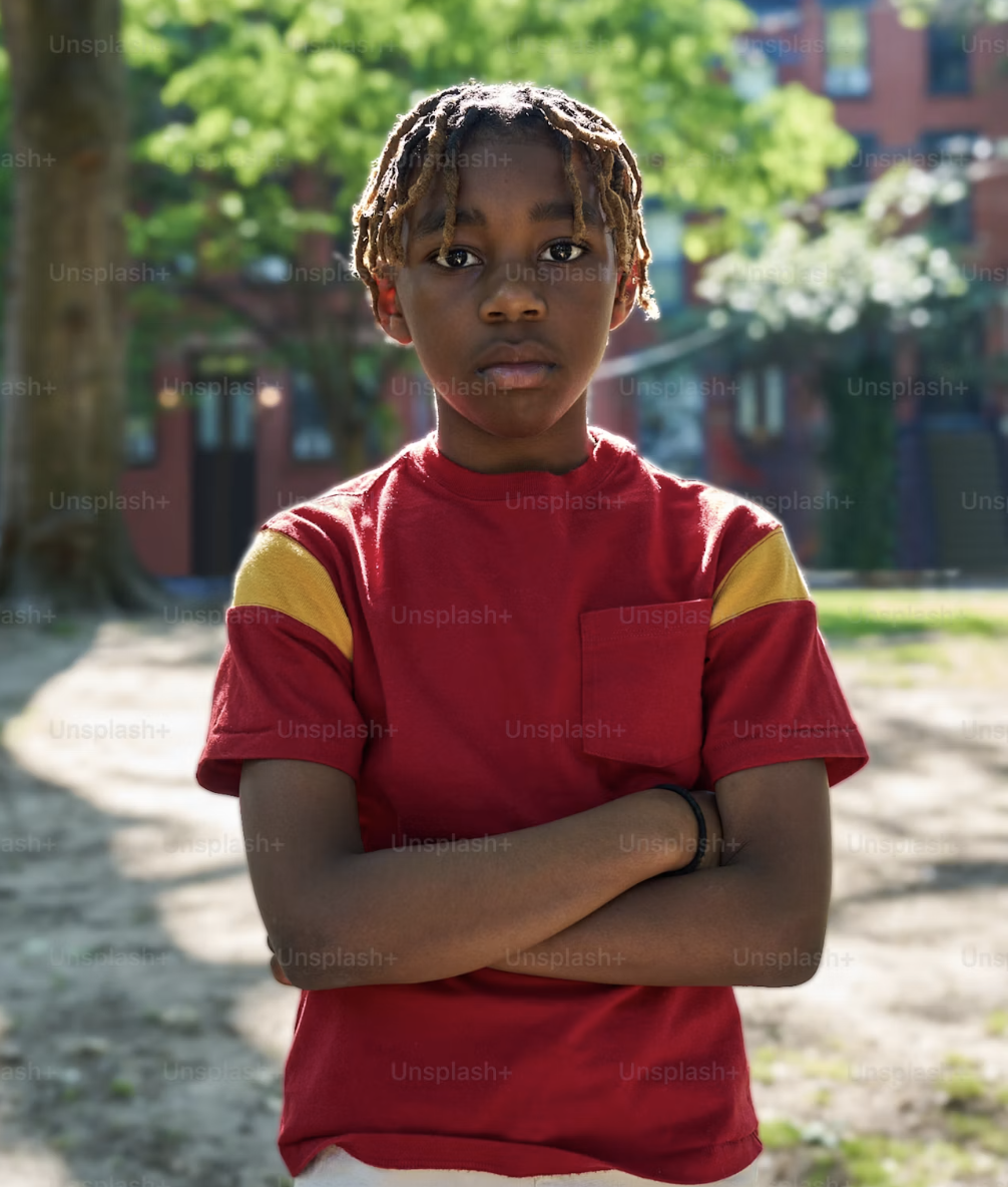 A young boy stands with his arms folded on a tree-lined neighborhood street